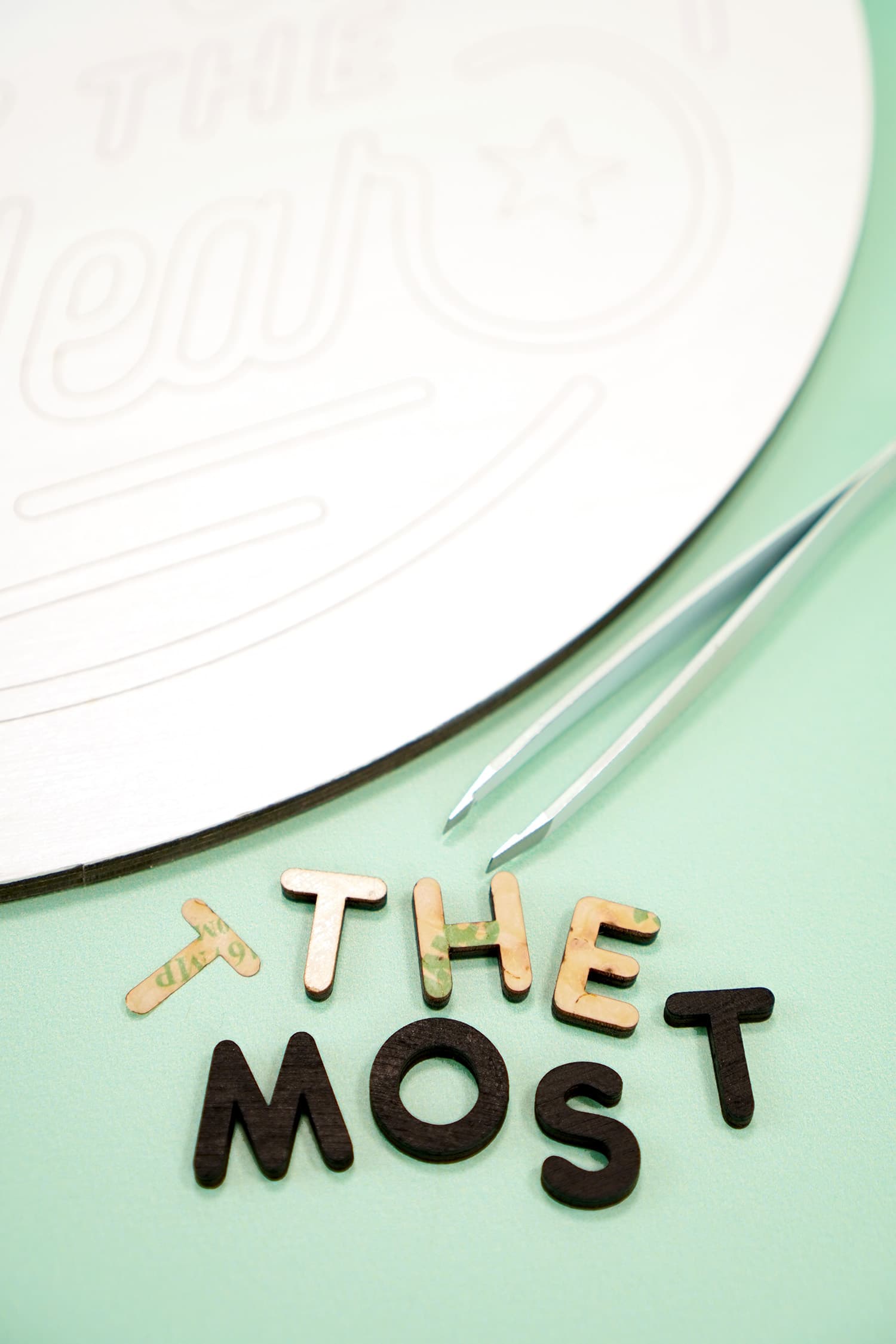 Close up of "The Most" laser cut wood sign pieces and tweezers on mint green background