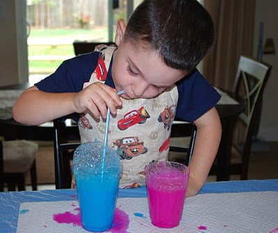 child blowing bubbles into cup of blue paint