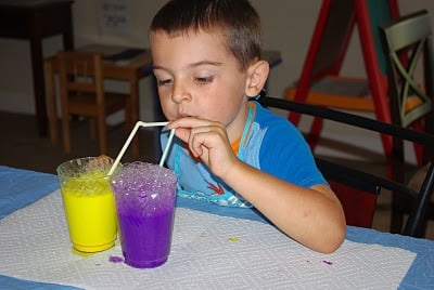 child blowing bubbles into cup of purple paint