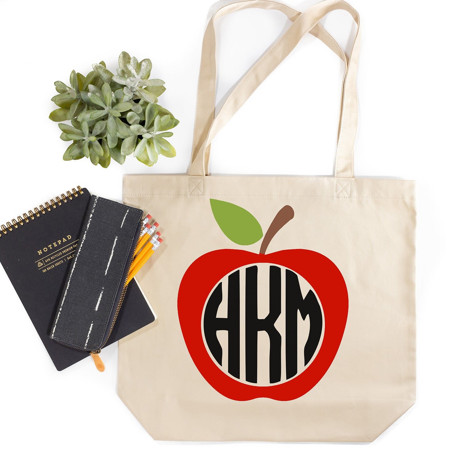 Tote bag with apple monogram design and notepad, pencils, and plant on white background