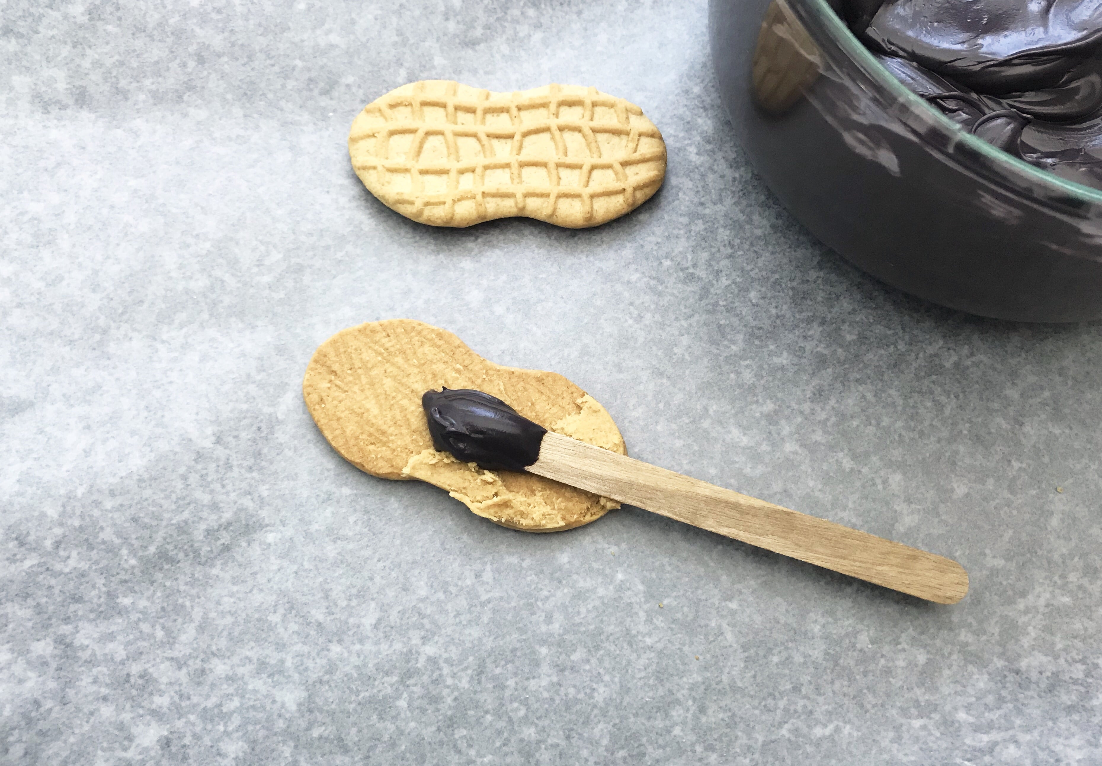 Icing on a popsicle stick being placed on a nutter butter cookie on a grey background