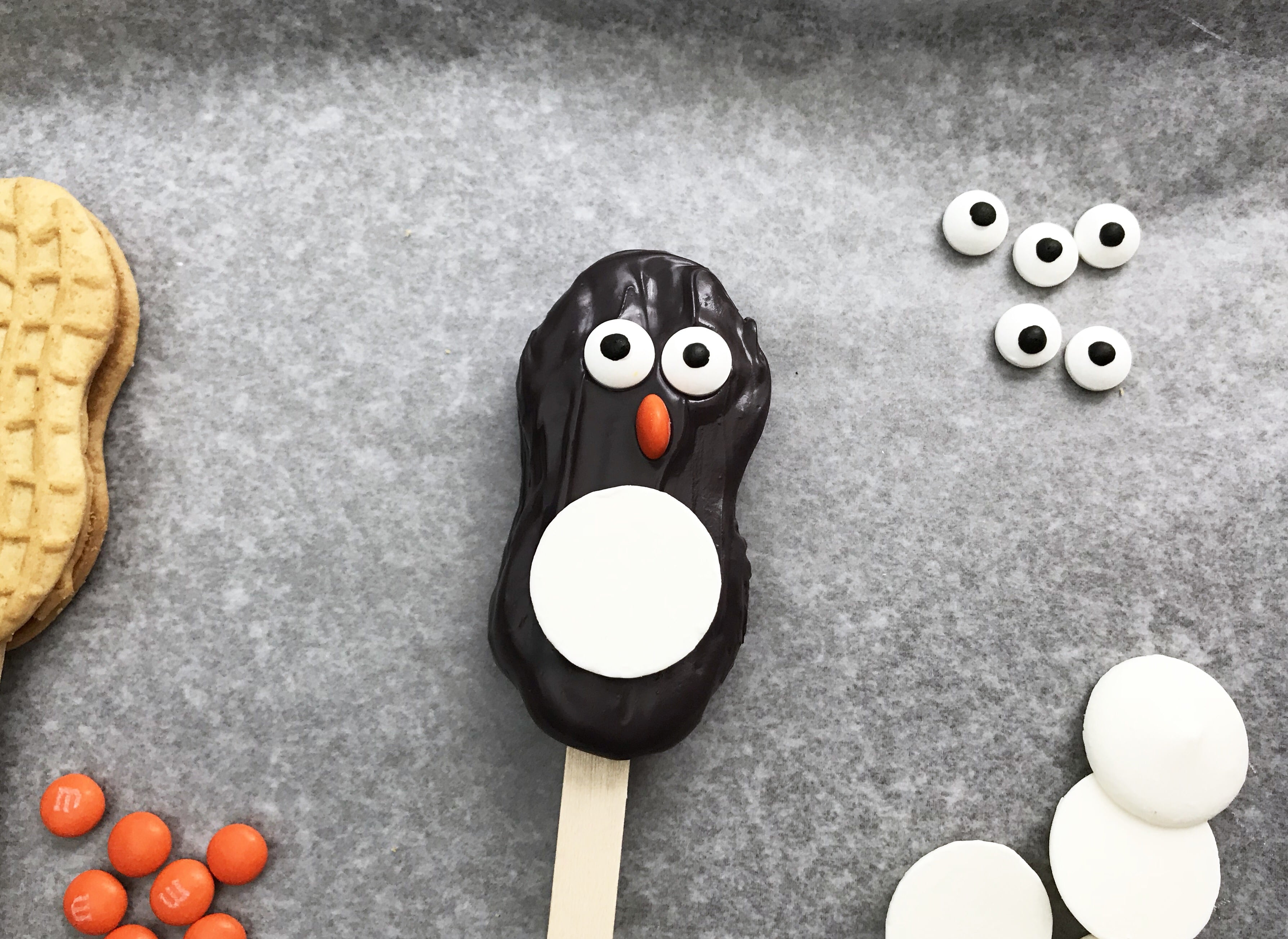 black icing covered nutter butter cookie pop with google eyes, candy beak on gray background