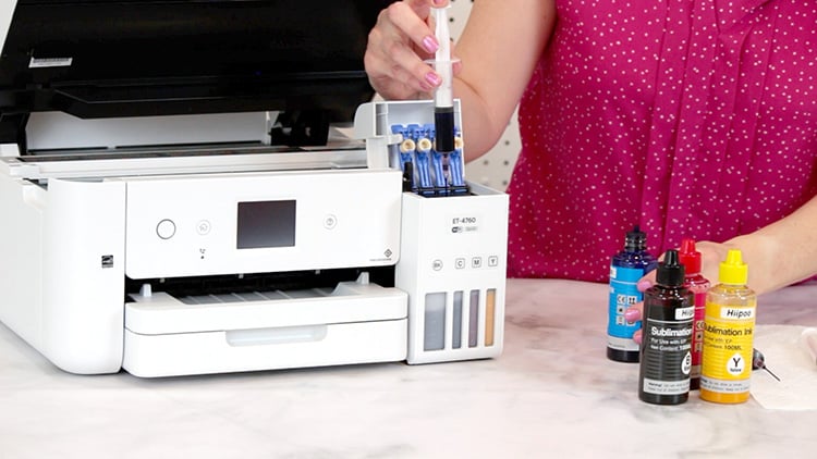 Hands filling an Epson EcoTank printer tank with sublimation ink from a syringe