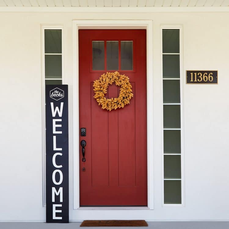 Black and white welcome sign beside a red front door with wreath