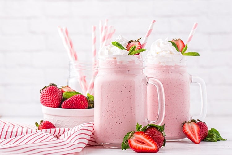 Two strawberry milkshakes in glass mugs with pink striped straws and strawberries