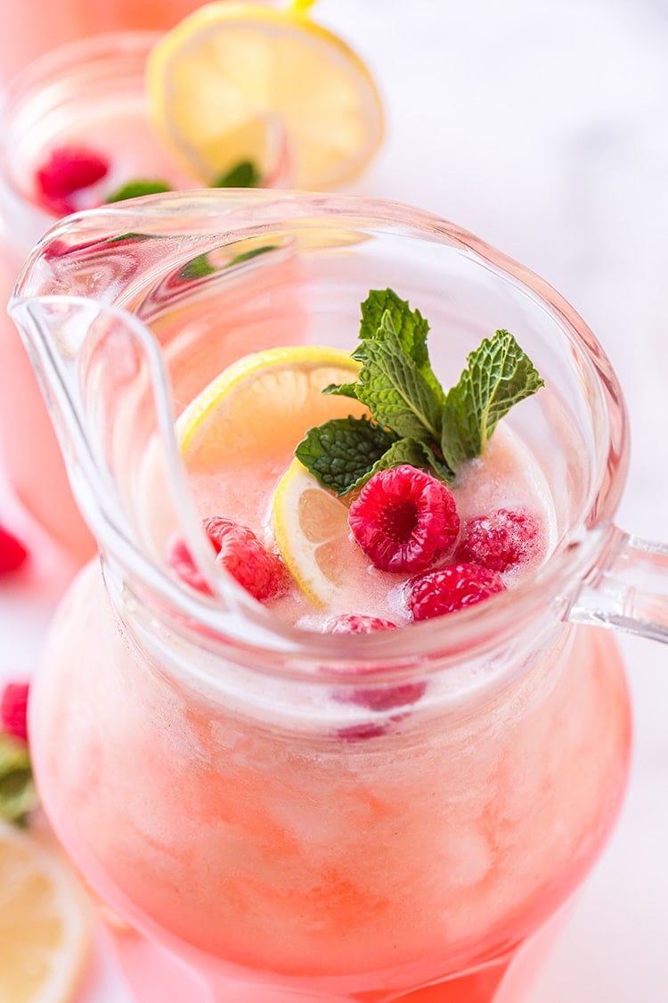 Close up view of raspberry lemonade in pitcher with lemon slices, fresh raspberries, and a mint sprig garnish