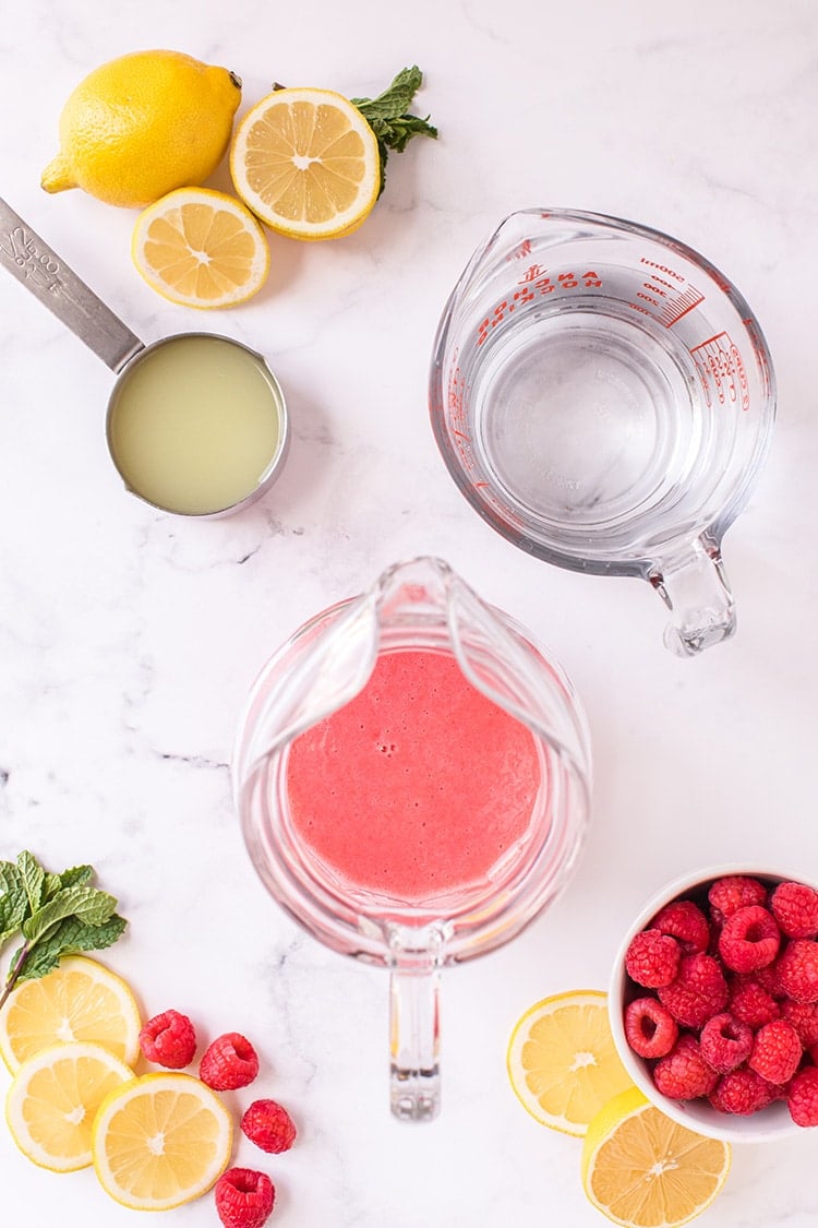 Overhead view of raspberry puree in pitcher surrounded by lemons and raspberries on a marble background