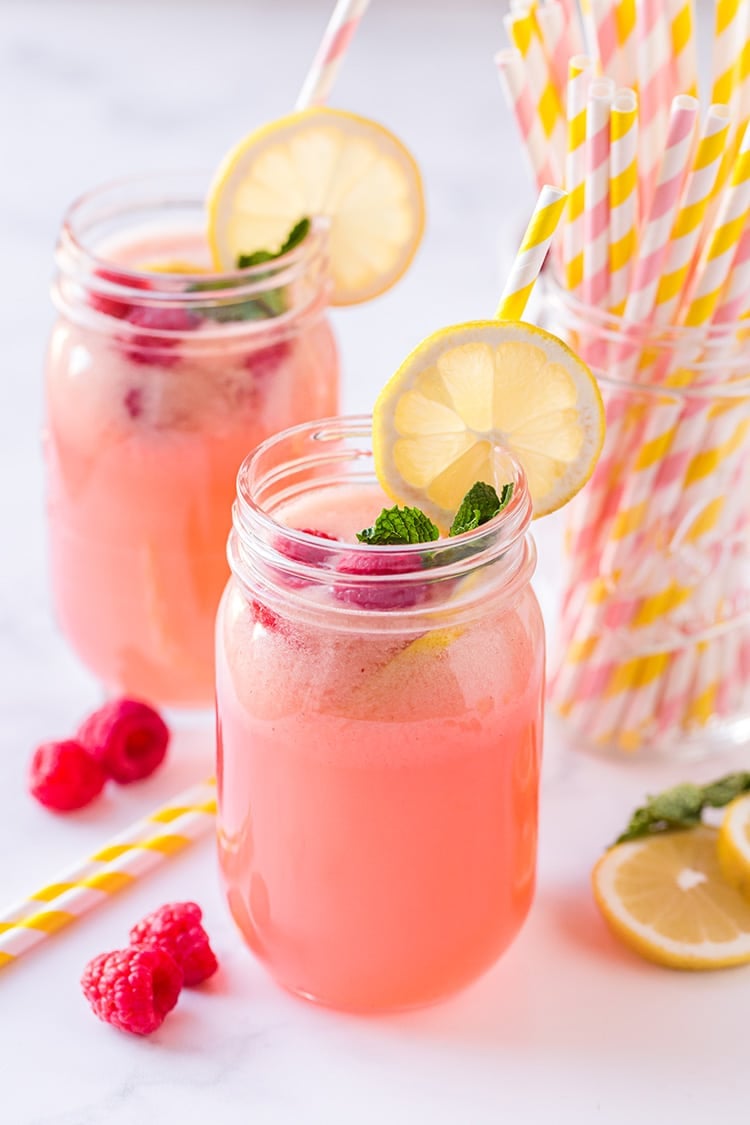 Glass of raspberry lemonade with pink and yellow striped straws, lemons, raspberries, and fresh mint garnish