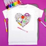 Partially completed coloring shirt with back to school themed design surrounded by fabric markers on purple background