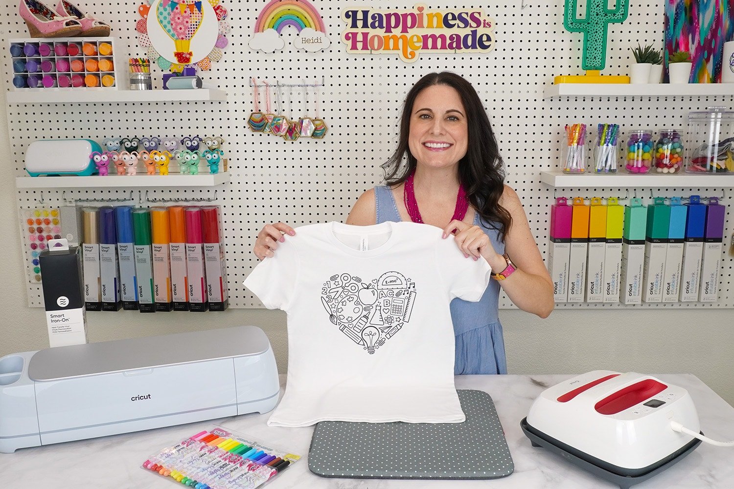 Heidi Holding Up Back to School Shirt in front of craft room pegboard of colorful supplies