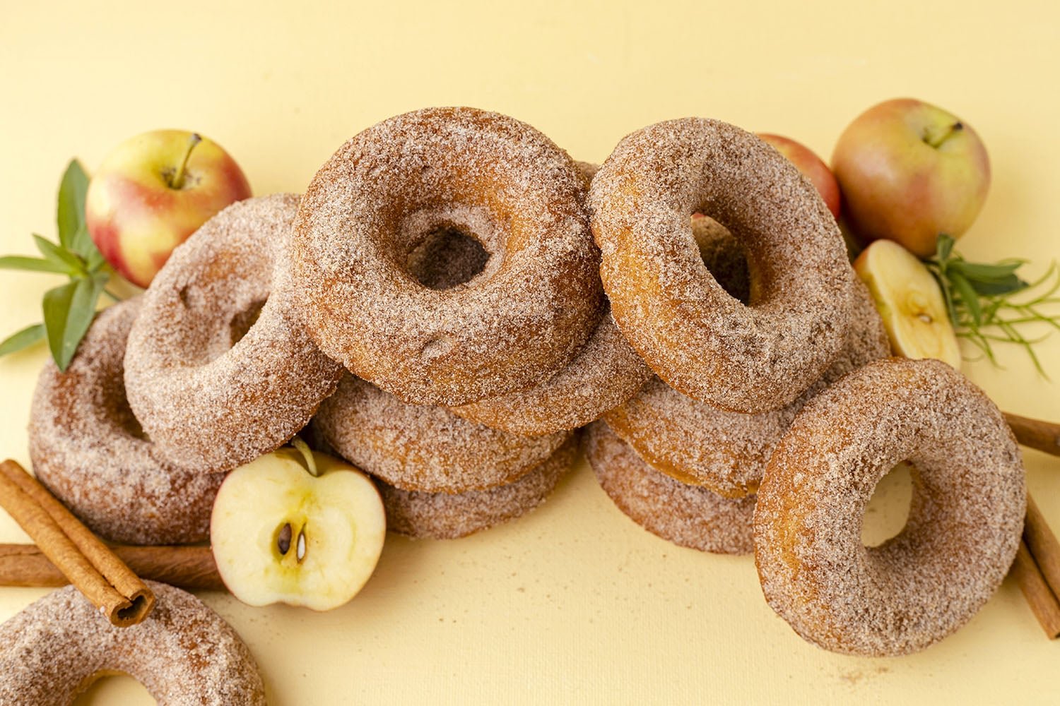 apple cider donuts in a stack with apples and cinnamon sticks on a yellow background