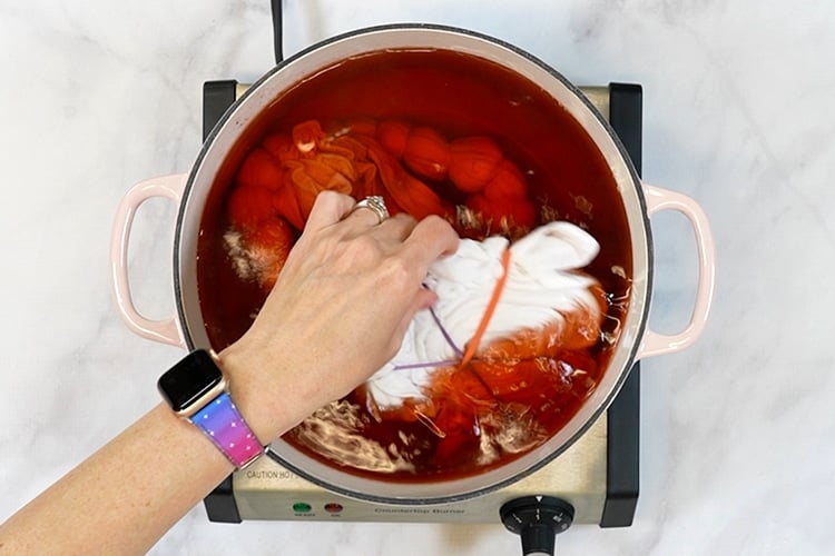 Hand adding a tie-dye folded shirt to a pot of red avocado pit dye