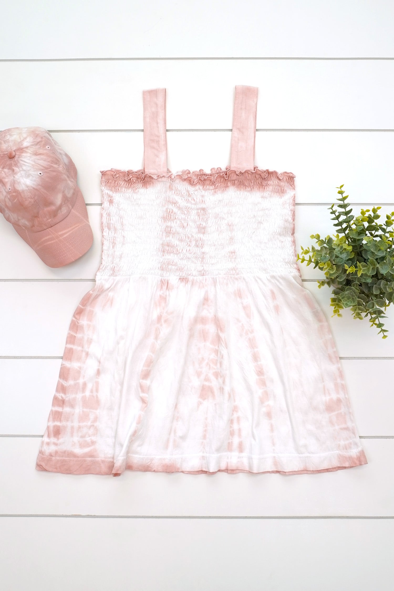 Blush pink tie-dye tank top and baseball hat made with avocado dye with plant on a white background