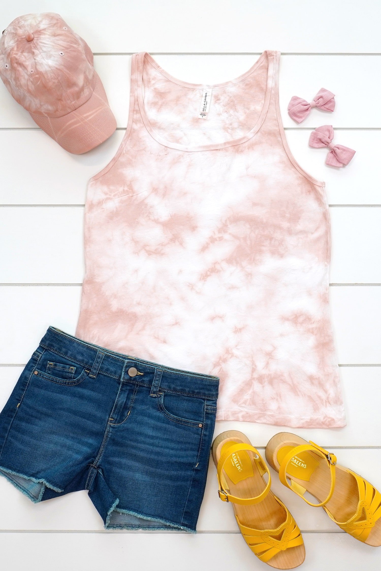 Blush pink tie-dye tank top and baseball hat made with avocado dye staged with an outfit and flowers on a white background