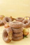 stack of apple cider donuts on yellow background with apples and cinnamon sticks