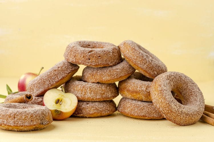 pile of apple cinnamon donuts on a yellow background with apples and cinnamon sticks