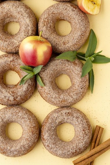 six apple cider donuts on a yellow background with apples, leaves, and cinnamon sticks