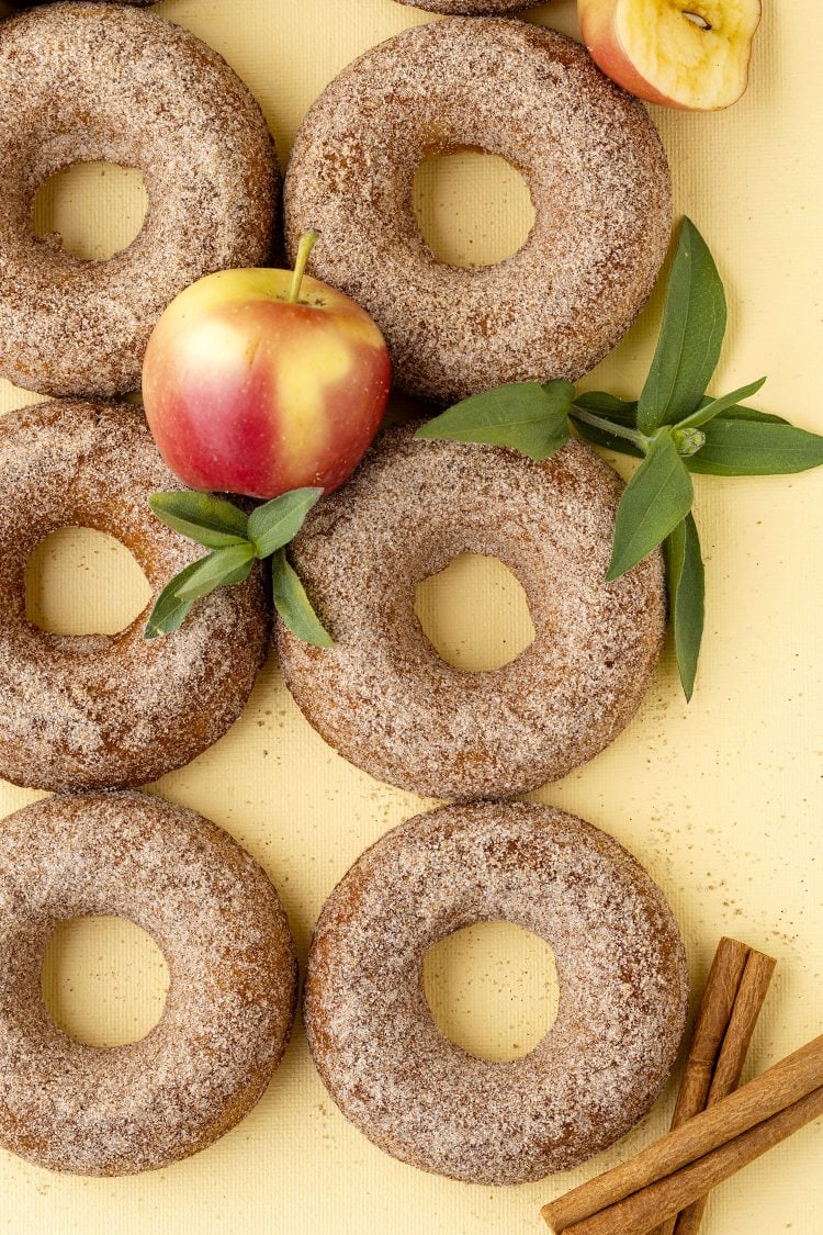 six apple cider donuts on a yellow background with apples, leaves, and cinnamon sticks