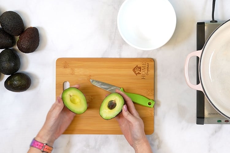 Hands holding a cut open avocado above cutting board with knife and pile of avocados