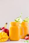 Two mango smoothies in mason jar glasses with lime wheels, mint leaves, and striped straws and sliced mangoes in foreground
