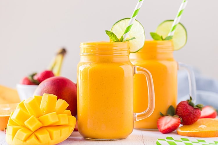 Two mango smoothies in mason jar glasses with lime wheels, mint leaves, and striped straws