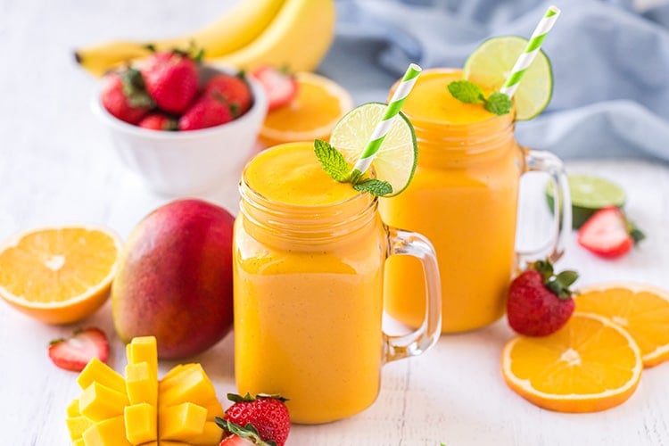 Two mango smoothies in mason jar glasses with strawberries and mangoes in the background