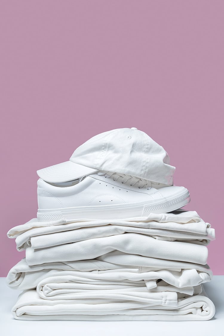 Stack of new white clothing, shoes, and hat on dusty purple background
