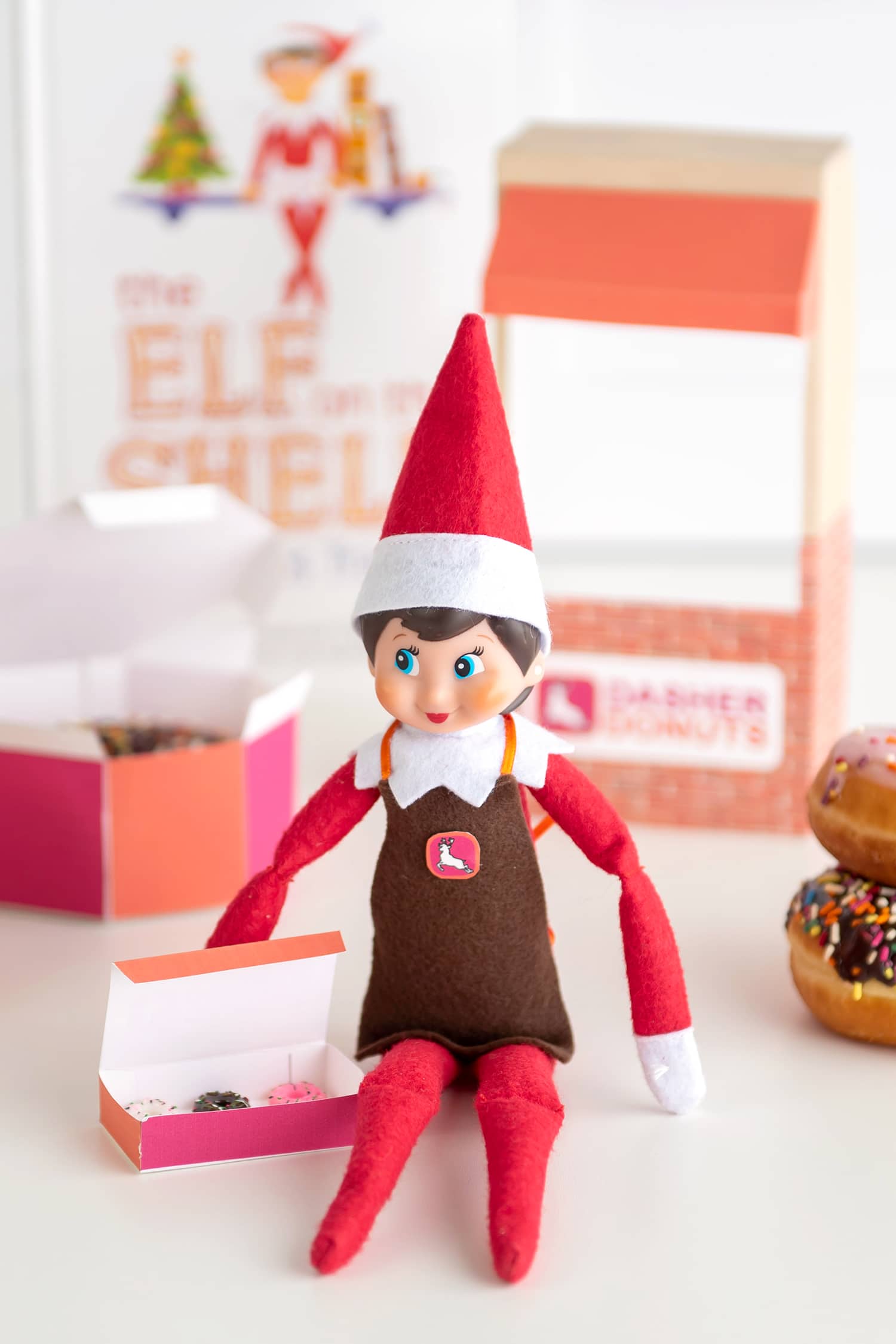 Girl Elf on the Shelf doll wearing an apron and sitting with a box of faux mini donuts