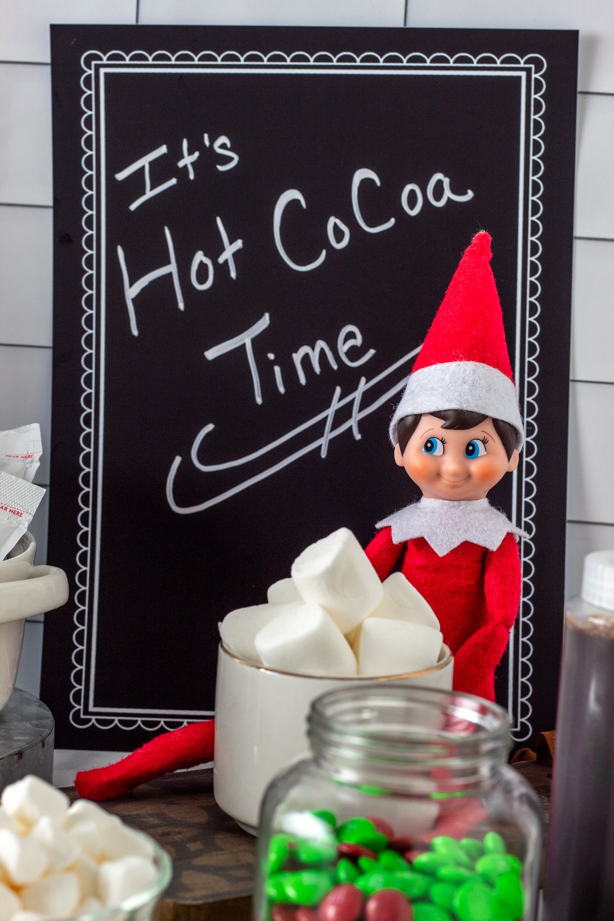 various hot cocoa bar items on kitchen counter with Elf on the Shelf and "It's Hot Cocoa Time" sign