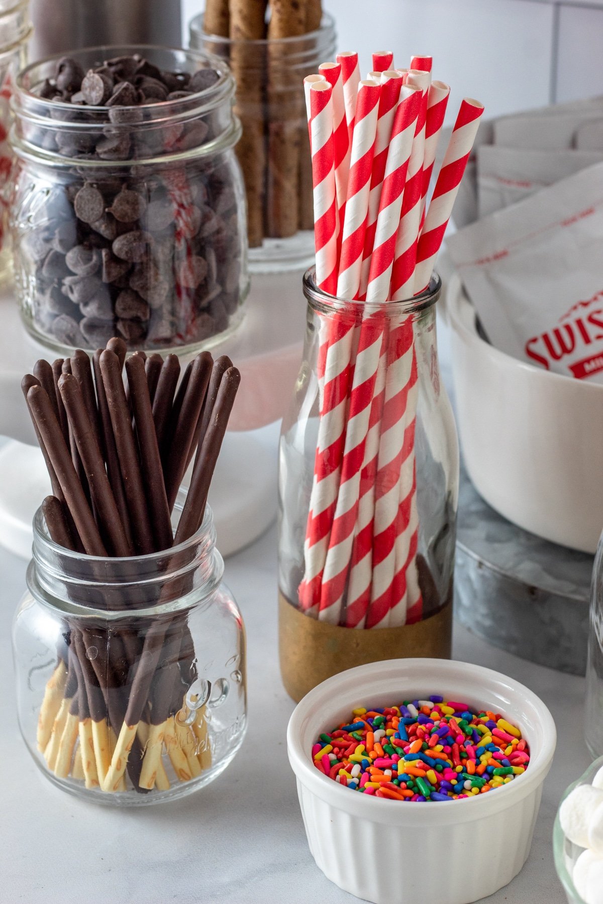 Pocky sticks, red striped straws, sprinkles, and chocolate chips in glass jars