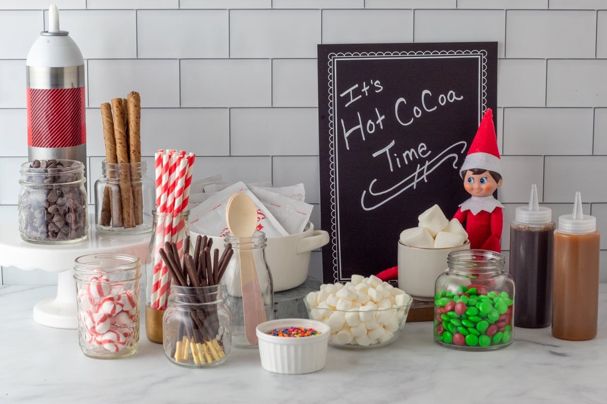 various hot cocoa bar items on kitchen counter with Elf on the Shelf