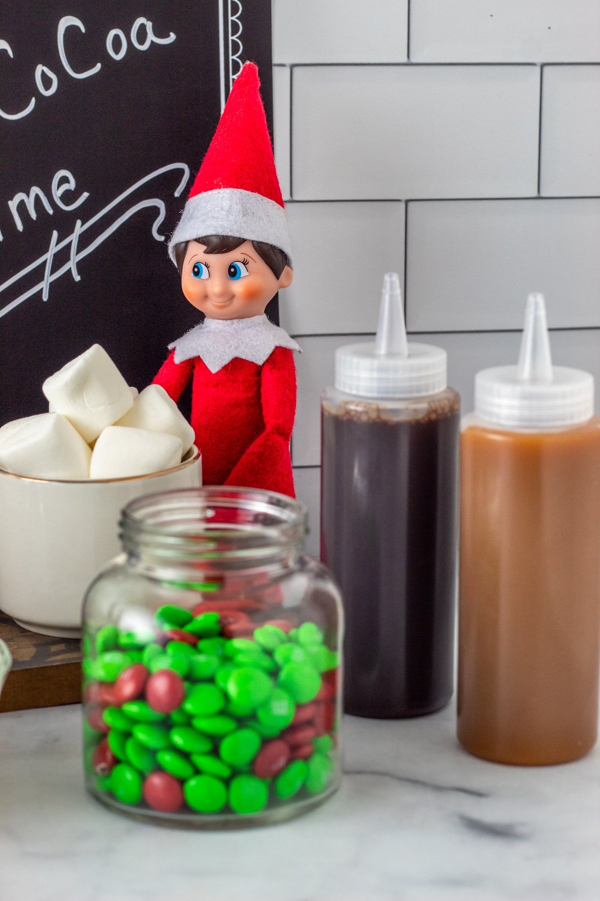 Chocolate and caramel syrups in bottles with Elf on the Shelf in hot cocoa bar