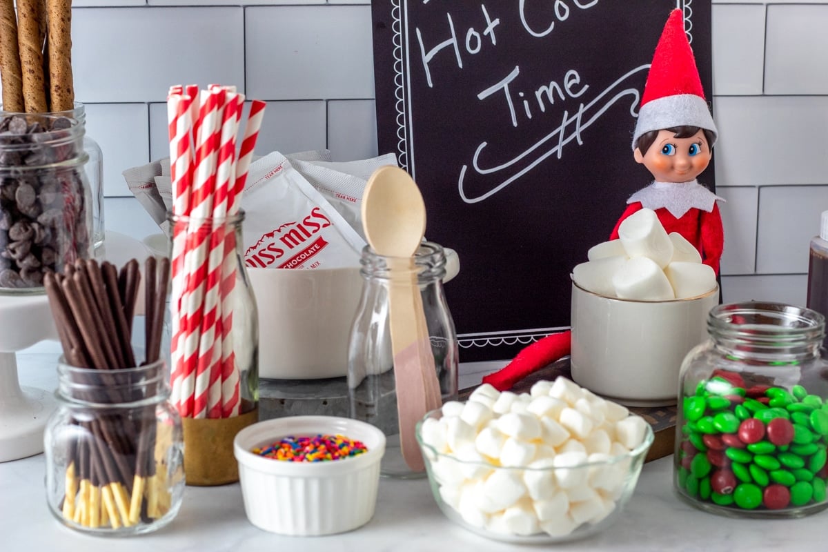 assorted hot cocoa bar toppings on kitchen counter with Elf on the Shelf doll