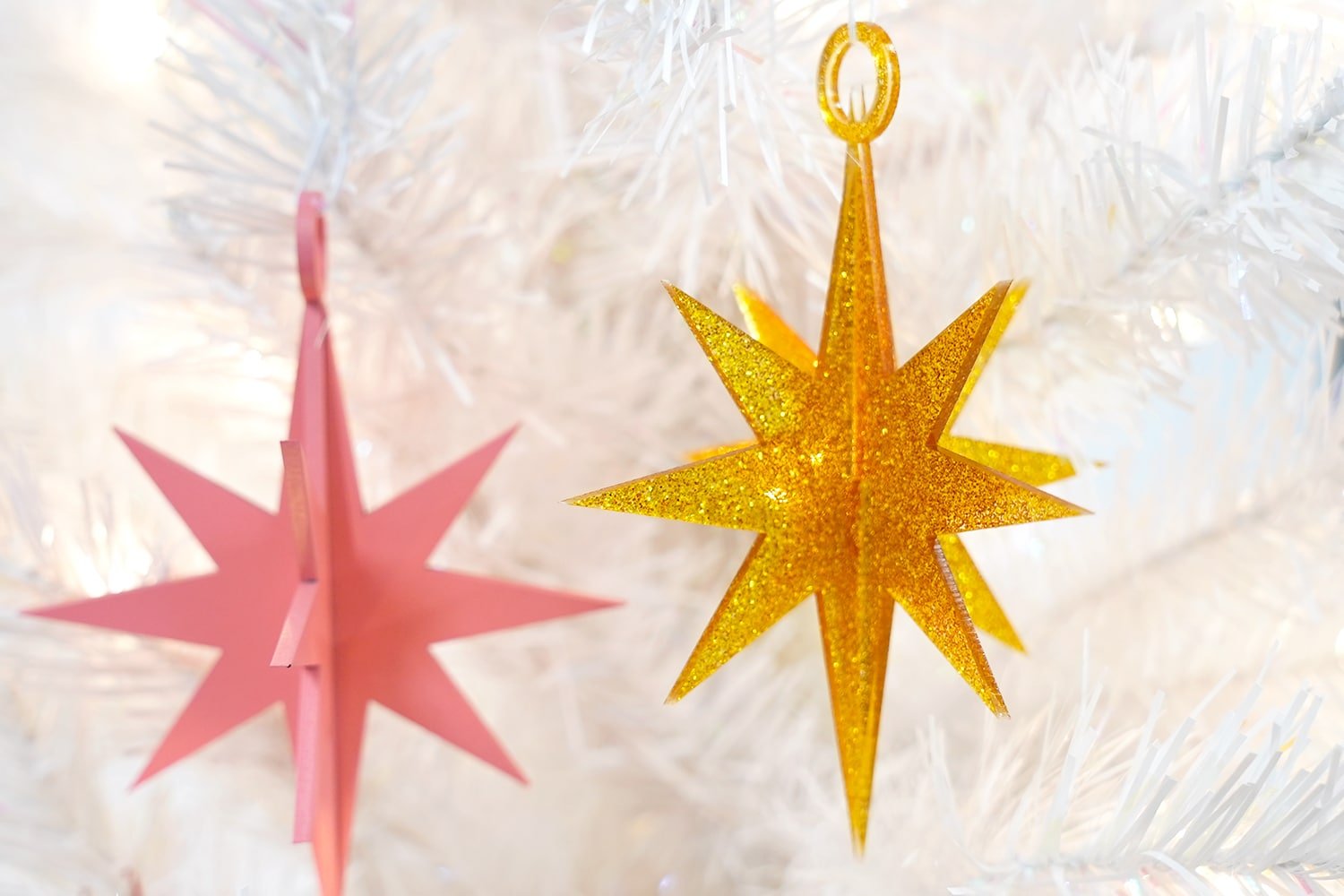 Gold glitter acrylic 3D star ornament on a white Christmas tree with pink star in background