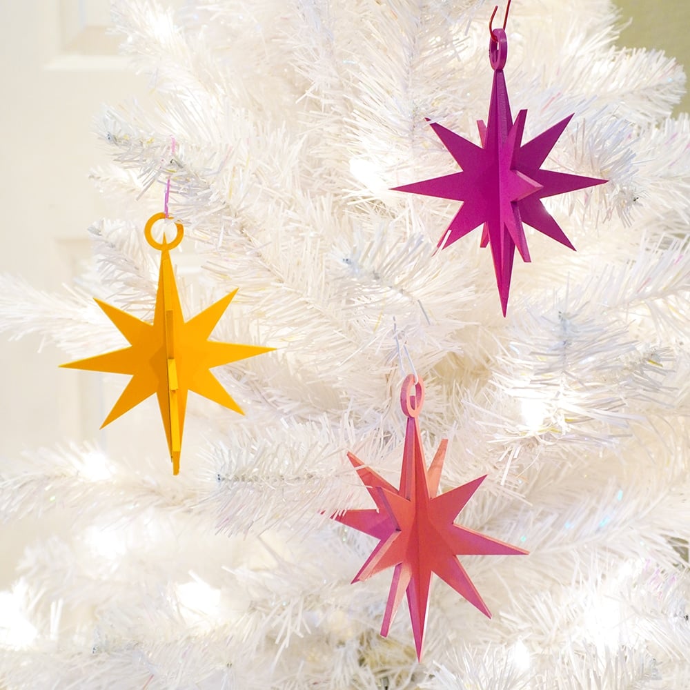Close up of white Christmas tree branches with three 3D star ornaments on them in pink, yellow, and magenta