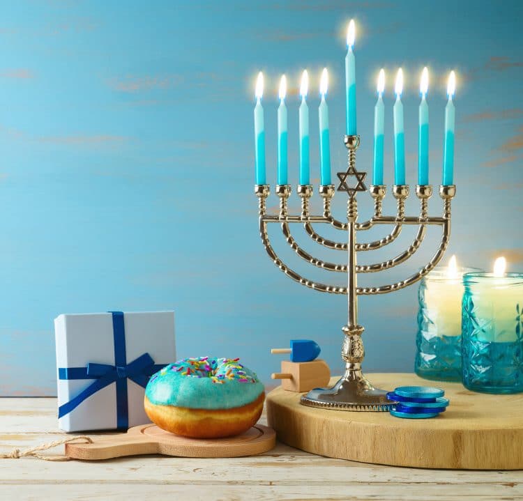 Fully lit menorah with teal blue candles, blue frosted donut, dreidels, and Hanukkah gifts