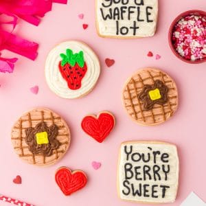 Waffle, strawberry, and heart sugar cookies with "I like you a waffle lot" and "You're berry sweet" cookies on pink background