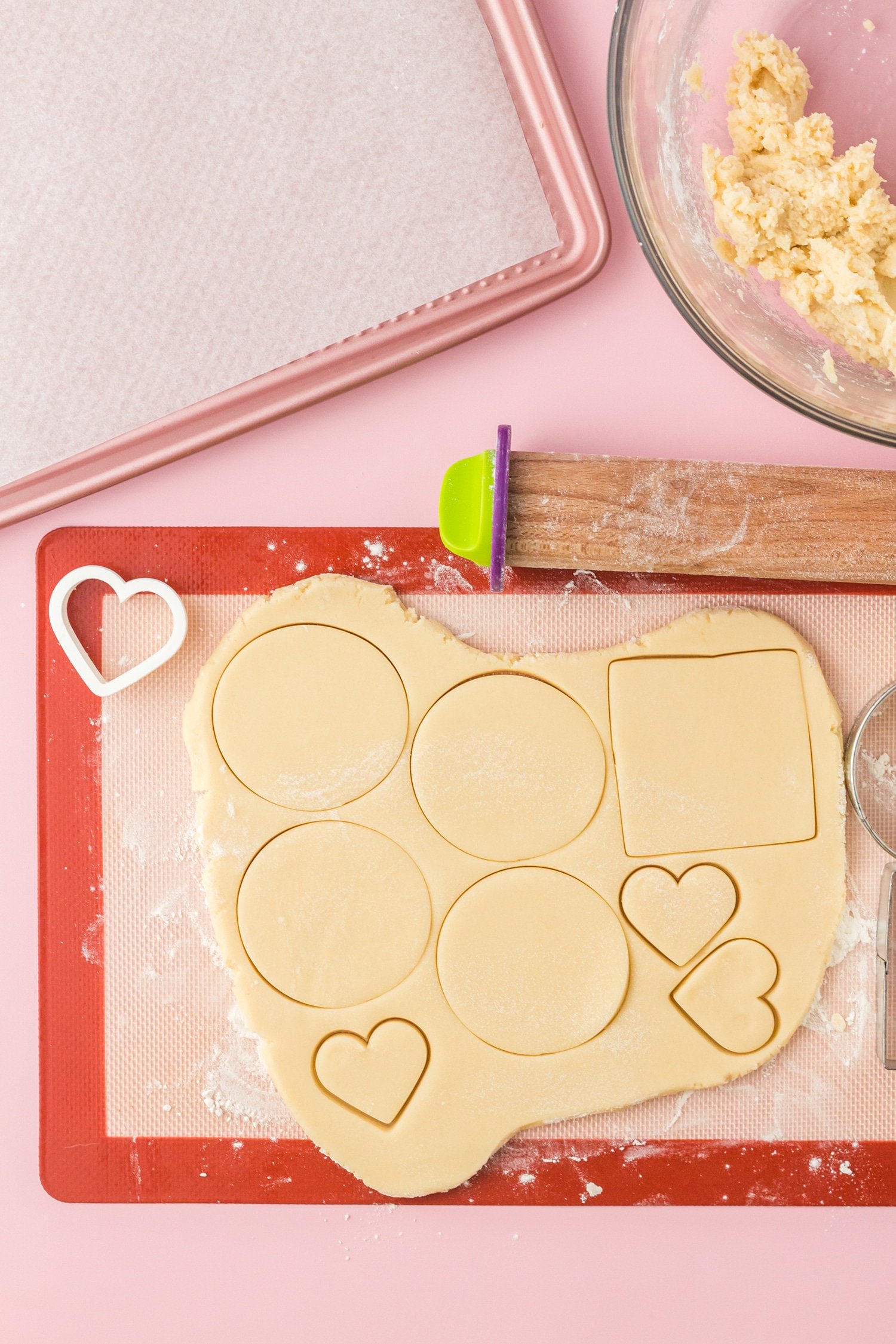 Sugar cookie dough and cutouts on a silicone mat on pink background