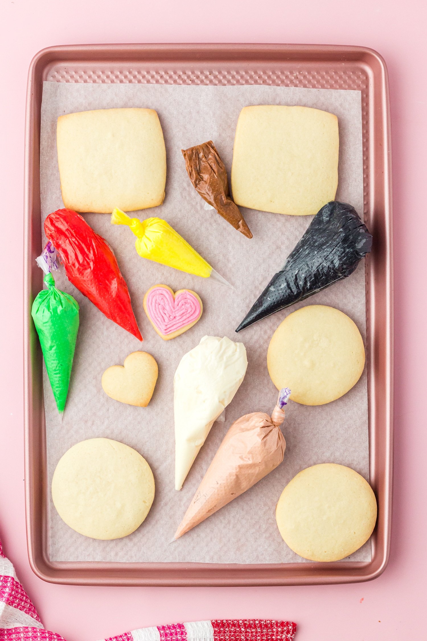 Colorful piping bags and baked sugar cookies on a pink baking sheet