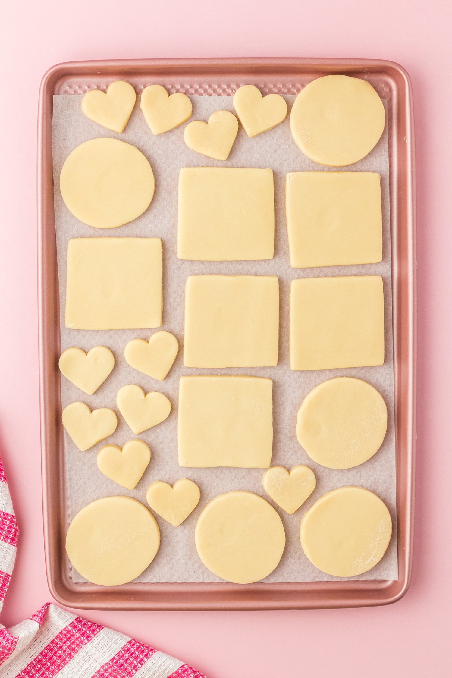 unbaked sugar cookie dough in heart, circle, and square shapes on pink baking sheet