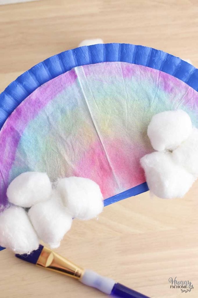 Pastel dyed coffee filter in rainbow shape with cotton ball clouds and purple paint brush.