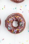 One chocolate donut with chocolate icing and rainbow sprinkles on white wood background