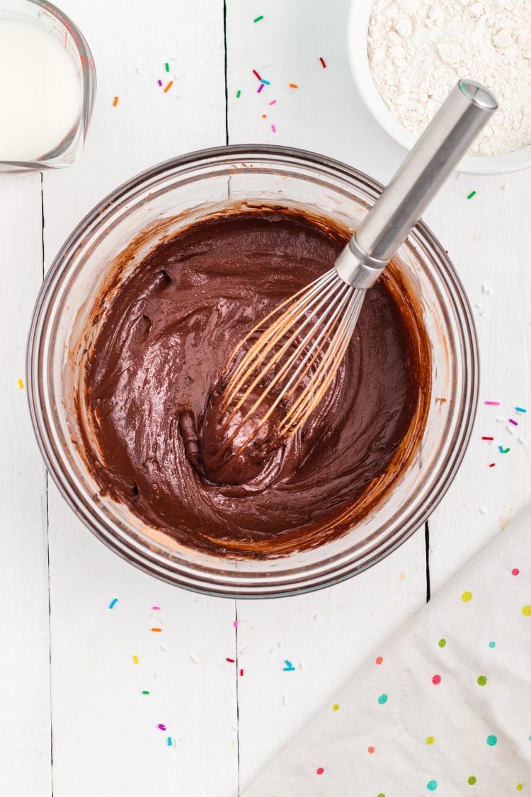 chocolate donut batter in mixing bowl with wire whisk on white background with sprinkles