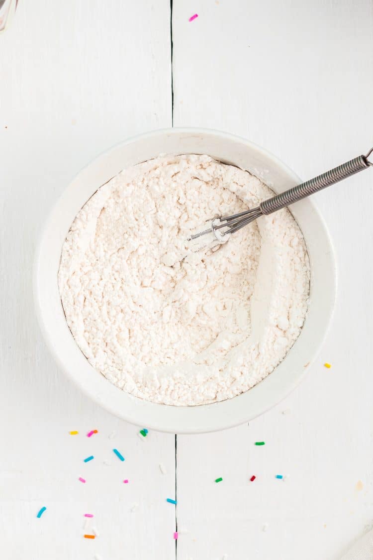 White flour in mixing bowl with whisk on white wood background