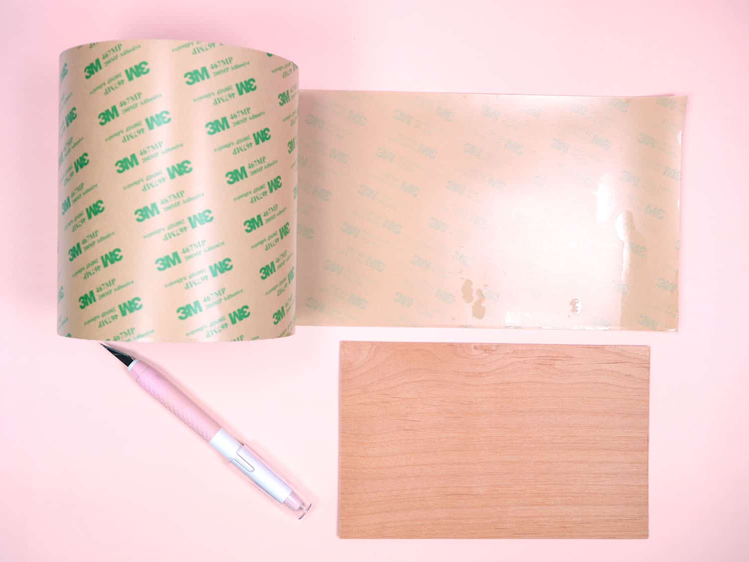 Partially unrolled roll of 3M tape, sheet of wood veneer, and craft knife on pink background