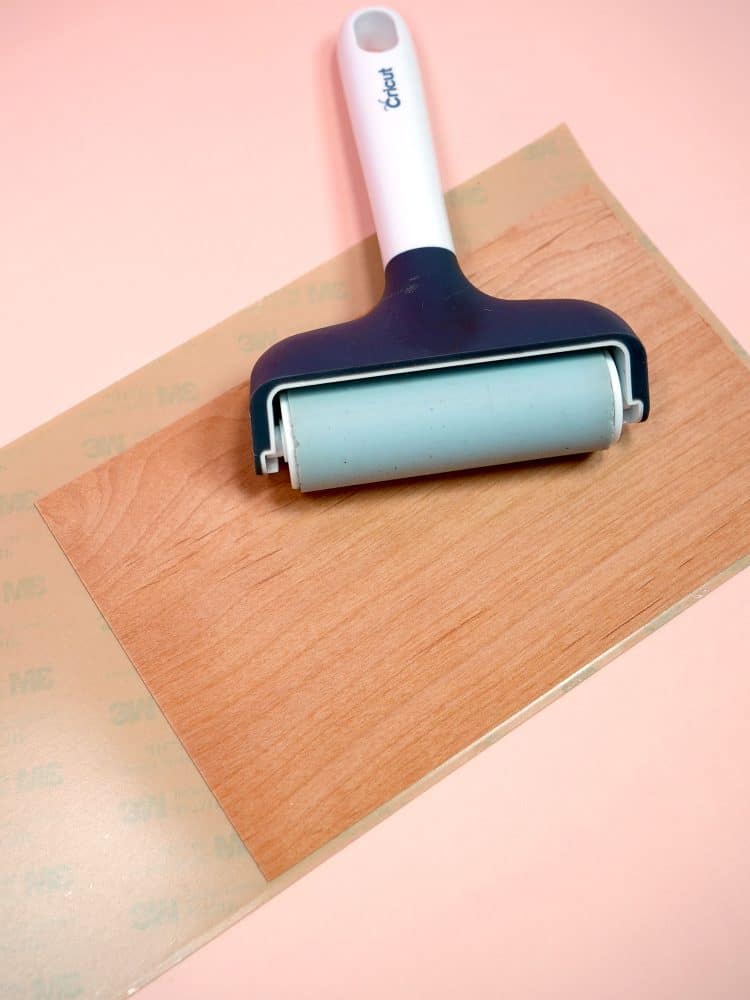 Using a brayer to apply 3M tape to a sheet of wood veneer on a peach background