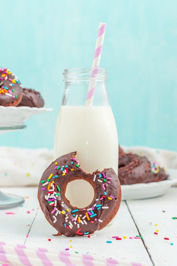 Chocolate donut with a missing bite in front of a small jar of milk with a striped straw on blue background