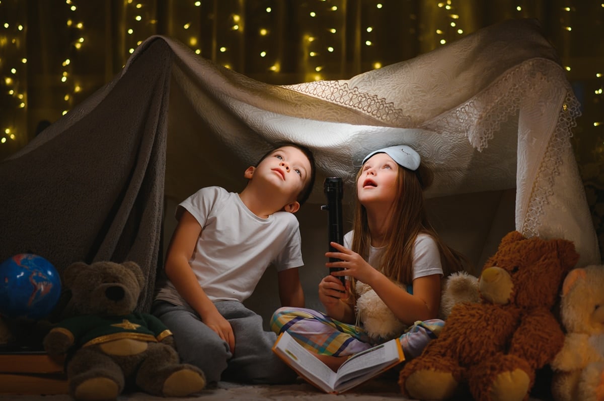 Two little children play at home in a blanket fort and flashlight 
