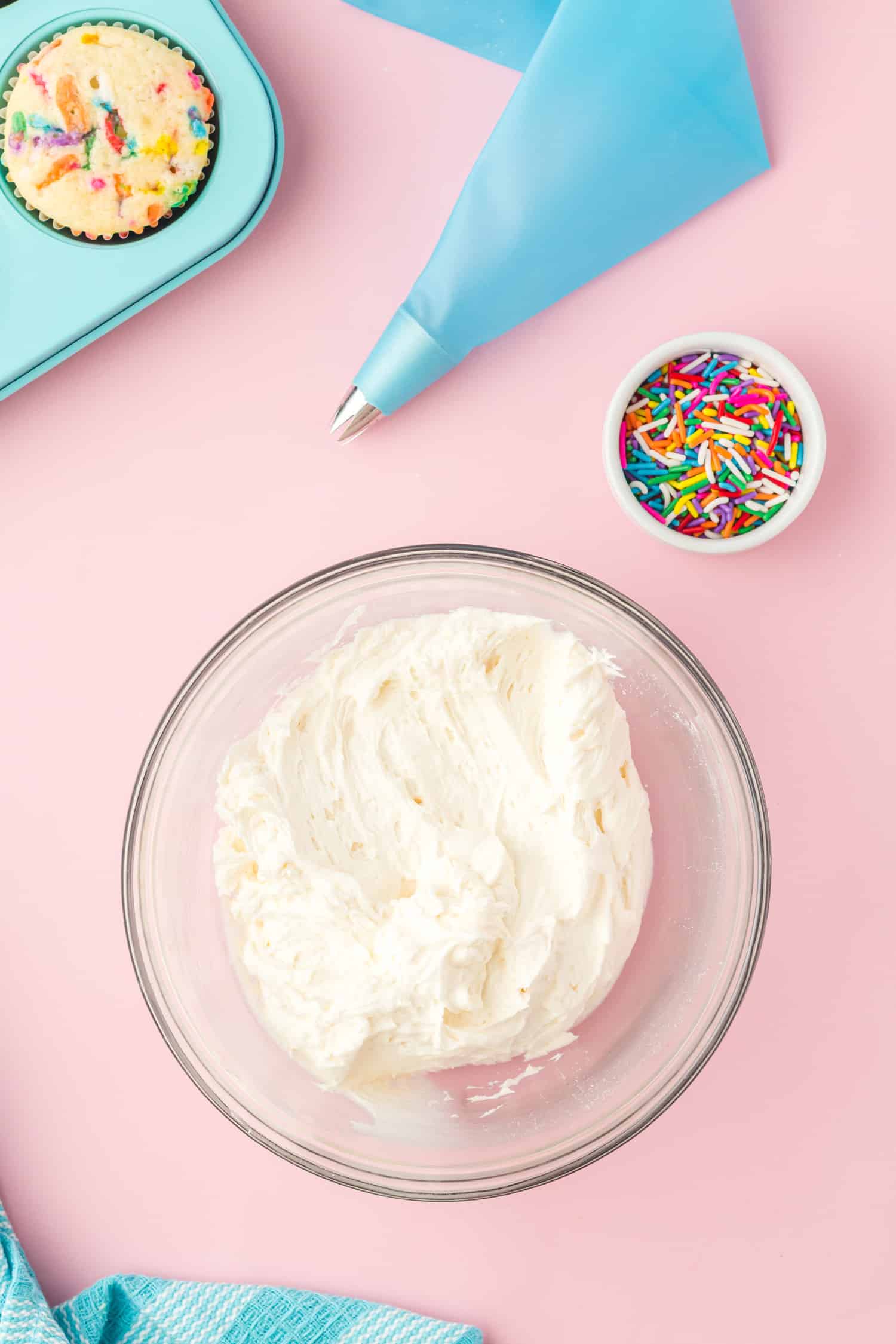 Buttercream icing in a glass bowl on pink background with sprinkles and unfilled icing bag