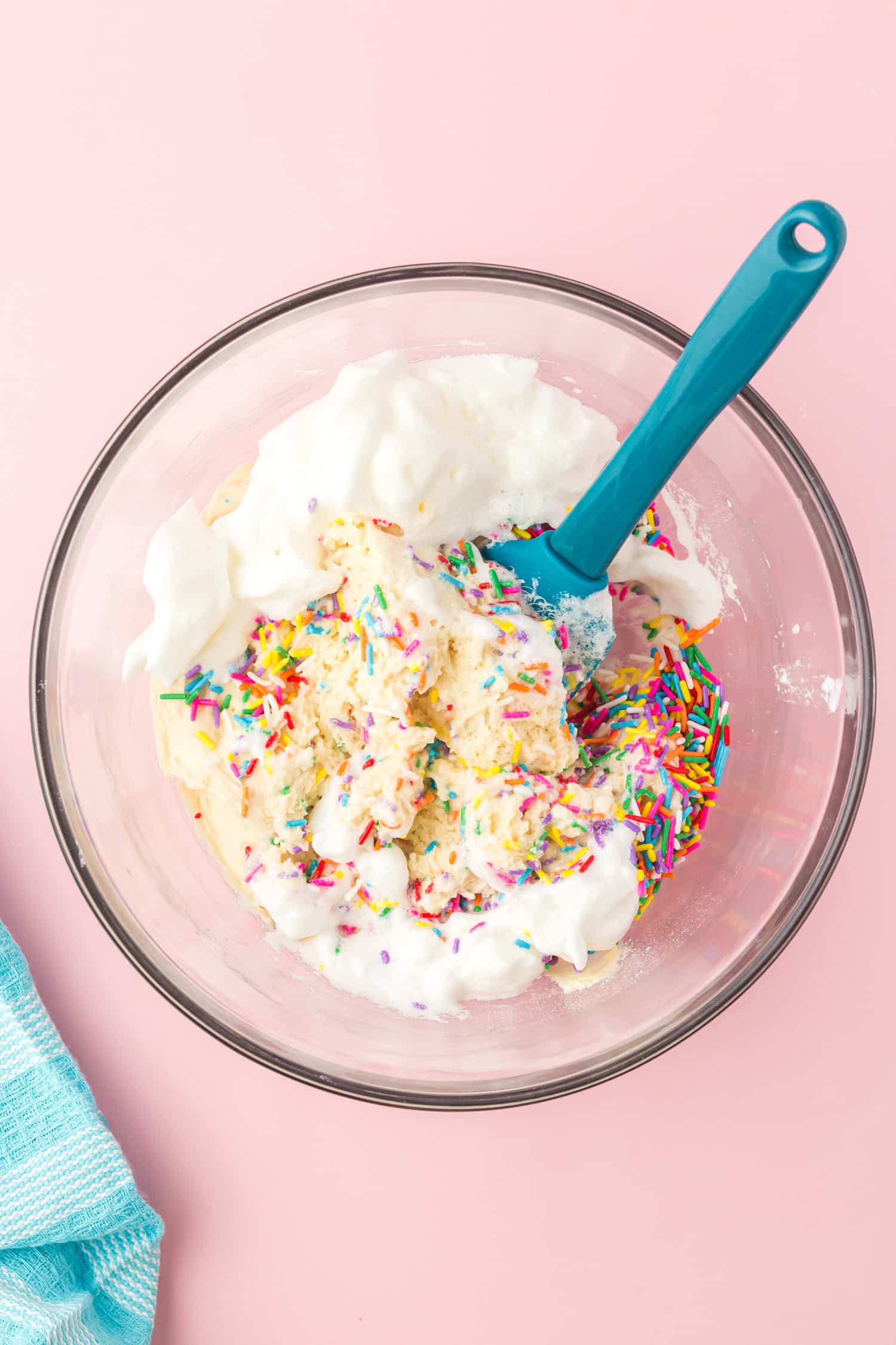 Glass bowl of whipped egg whites being folded into Funfetti cupcake batter with teal blue spatula